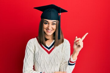 Young brunette girl wearing graduation cap smiling happy pointing with hand and finger to the side