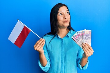 Poster - Young latin woman holding poland flag and zloty banknotes smiling looking to the side and staring away thinking.