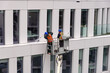 Two workers wearing safety harness wash office building facade at height standing in a crane cradle or aerial platform using pressure washer and mops