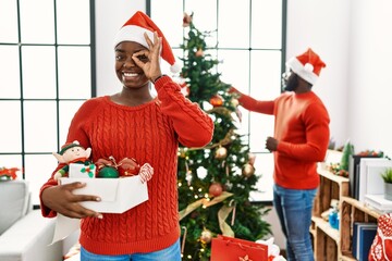 Young african american couple standing by christmas tree doing ok gesture with hand smiling, eye looking through fingers with happy face.