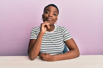 Poster - Young african american woman wearing casual clothes sitting on the table looking confident at the camera with smile with crossed arms and hand raised on chin. thinking positive.