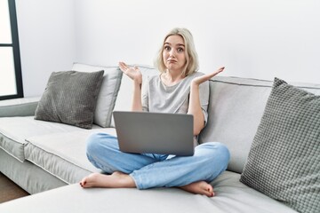 Poster - Young caucasian woman using laptop at home sitting on the sofa clueless and confused expression with arms and hands raised. doubt concept.