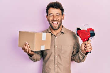 Poster - Handsome man with beard holding packing tape holding cardboard smiling and laughing hard out loud because funny crazy joke.