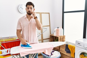 Poster - Young man with beard ironing clothes at home mouth and lips shut as zip with fingers. secret and silent, taboo talking