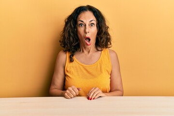 Poster - Beautiful middle age woman wearing casual clothes sitting on the table afraid and shocked with surprise and amazed expression, fear and excited face.