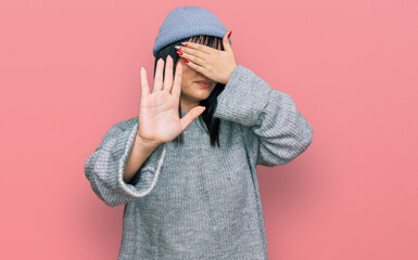 Poster - Young hispanic woman wearing cute wool cap covering eyes with hands and doing stop gesture with sad and fear expression. embarrassed and negative concept.