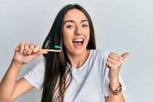Young Hispanic Girl Holding Toothbrush With Toothpaste Pointing Thumb Up To The Side Smiling Happy With Open Mouth