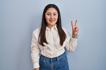 Wall Mural - Young latin woman standing over blue background showing and pointing up with fingers number two while smiling confident and happy.