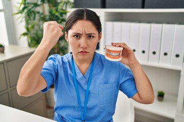 Canvas Print - Young latin dentist woman holding denture with braces annoyed and frustrated shouting with anger, yelling crazy with anger and hand raised