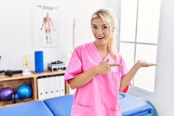 Poster - Young caucasian woman working at pain recovery clinic amazed and smiling to the camera while presenting with hand and pointing with finger.