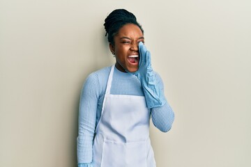 Poster - African american woman with braided hair wearing cleaner apron and gloves shouting and screaming loud to side with hand on mouth. communication concept.