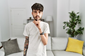 Poster - Hispanic man with beard at the living room at home asking to be quiet with finger on lips. silence and secret concept.