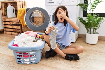 Sticker - Young hispanic girl doing laundry holding socks peeking in shock covering face and eyes with hand, looking through fingers with embarrassed expression.