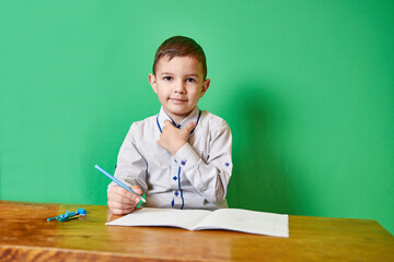 Cute smiling boy holding a pen in his hand sitting at a table with an open notebook on a green background.