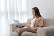 © Puwasit Inyavileart - Asian woman sitting on the sofa with a laptop, Looking at laptop screen, Relaxing at home, Working through the Internet communication system, Happy lifestyle, Feel good.