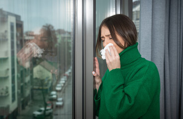 Young sick unhappy woman suffering from common cold or flu standing near window looking out wipes her nose with tissue. Self isolated ill female stay at home until she recover and get well.