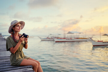 Wall Mural - Photography and travel. Young woman in hat holding camera sitting on wooden fishing pier with beautiful tropical sea view.