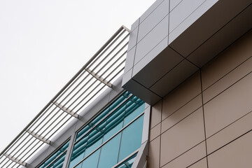 Wall Mural - The upward view of a modern commercial building with beige and grey metal composite panels, blue glass windows, and a slated grate on the edge of the roof. The sky is cloudy and white in color.