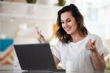 Fototapeta Pomosty - smiling remote working woman chatting and waving hand infront of a laptop or notebook during a video conference on her work desk in her modern bright living room home office 