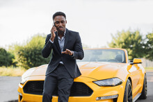 Portrait Of Rich African Businessman Wearing Suit, Standing Near His Luxury Yellow Cabriolet Car And Using Smartphone While Holding In Hand. Concept Of People, Technology And Transport.
