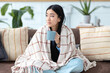 © kucherav - Asian young woman with a cup of coffee sitting on the couch at home covered with blanket feeling unwell, relaxation at home