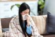 © kucherav - Ill Asian young woman sitting on the couch at home covered with blanket, blowing nose, got flu