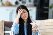 © kucherav - Ill Asian young woman sitting on the couch at home covered with blanket, blowing nose, feeling unwell, has a cold or flu