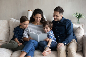 Happy family reading book together, relaxing on couch at home, smiling loving mother telling to father and adorable little son and daughter funny fairy tale story, spending leisure time weekend