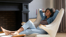 Full Length Peaceful Happy Millennial Generation African American Female Homeowner Resting With Folded Arms In Armchair With Legs On Footstool, Doing Yoga Breathing Exercises, Napping Alone At Home.
