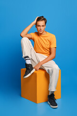 Wall Mural - Positive man in orange undershirt and white sweatpants sits on wooden cube over blue background