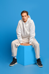Wall Mural - Cheerful man in white sweatshirt and white sweatpants sits on wooden cube over blue background