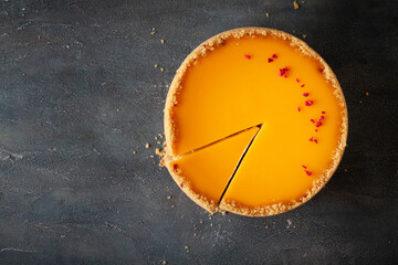 Wall Mural - Flat lay of cheesecake with yellow mango flavoured layer decorated with red crumbs of freeze-dried raspberries. Cake cut on the dark marble background