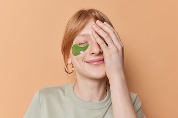 Pleased ginger freckled teenage girl covers half of face with palm keeps eyes closed undergoes beauty procedures applies green hydrogel patches isolated over brown background. Skin care concept