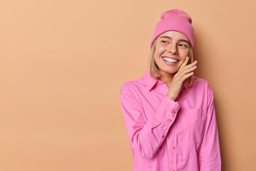 Wall Mural - Horizontal shot of happy young European woman touches face and smiles pleasantly concentrated away wears pink hat and shirt isolated over brown background blank copy space for your promotion