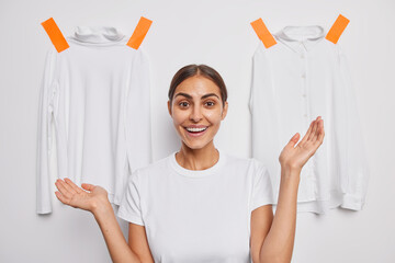 Wall Mural - Good looking cheerful woman with dark combed hair keeps palms raised over plastered clothing demonstrates outfit to wear smiles broadly isolated over white background. Advertisement concept.