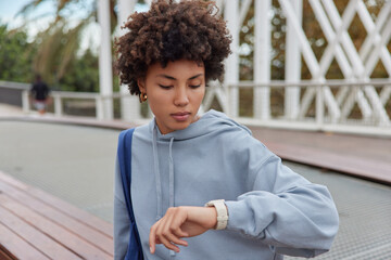Wall Mural - Outdoor shot of curly haired woman wears casual hoodie checks time on smartwatch carries bag waits for someone has serious expression waits for trainer to start sport training. People and lifestyle