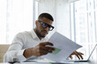 © fizkes - Serious African American manager in stylish glasses reading corporate document with note, sales report, boss order, doing paperwork. Legal expert checking document at workplace with laptop