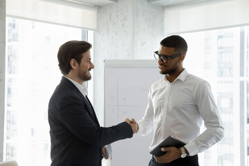 Wall Mural - Two diverse smiling business men shaking hands in office boardroom. Employee hiring candidate after job interview. Client thanking manager, lawyer with handshake for consultation, negotiation