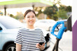 Happy Asian young woman using ev charging application on smartphone connect with the charging station.