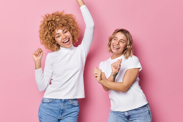 Wall Mural - People fun entertainment concept. Positive carefree women dance and sing together shake arms dressed in casual clothes move against pink background. Two glad girls move to music like nobody watching