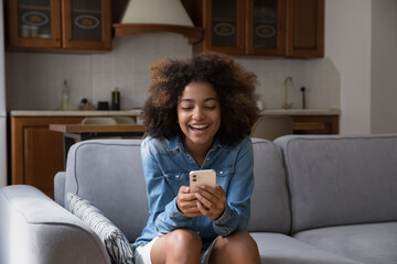 Cheerful happy gen Z teen girl receiving exciting message on smartphone, reading text in screen, smiling, laughing, sitting on couch, using online virtual app on mobile phone