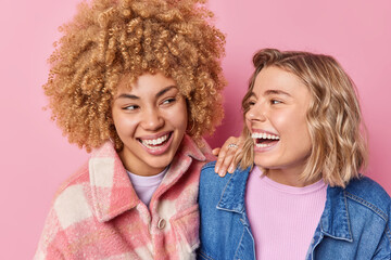 Wall Mural - Positive friendly women have fun laughs joyfully look at each other sped free time together dressed in fashionable clothes isolated over pink background. People frinendship emotions concept.