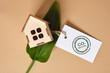 green leaves, miniature wooden house model and white tag on brown background