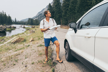 Young man having a problem with orientation on road during his journey with a car. Caucasian guy tourist looking on a map while standing next to car.