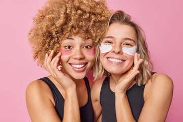 Canvas Print - Young happy women apply beauty pathes under eyes smile broadly enjoys skin care procedures wear casual black t shirts isolated over pink background. Two female models put on pads to remove wrinkles