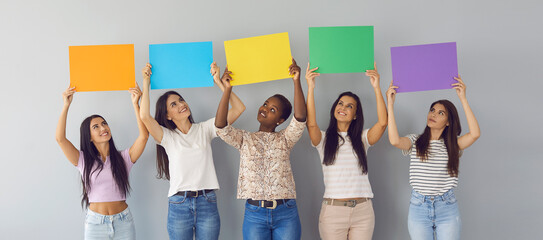 Banner background with group of happy young multiracial women looking up at colorful orange, blue, yellow, green, purple mockup paper signs they're holding standing together by white grey studio wall