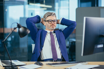 Wall Mural - pensive and dreamy experienced businessman owner boss, sitting at work, throwing his hands behind his head, resting