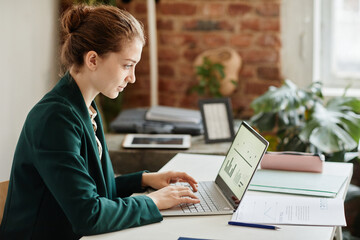 Young confident businesswoman in formal clothes typing on laptop keyboard while analyzing financial data on screen