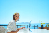 Fototapeta  - Beautiful young mixed race woman smiling to camera enjoying using laptop online internet at outdoor resort cafe with sea view