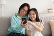 © Prostock-studio - Loving Asian grandmother and her cute little granddaughter using smartphone on couch at home